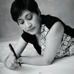 A head shot of Khairani Barokka. Black and white photo of an Indonesian woman with short hair, earrings, and a patterned dress, lying down on her front, pen in hand, ready to write. Picture credit: Derrick Kakembo.