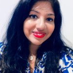 Image of S Niroshini. A south asian woman who smiles at the camera. She has long black hair and wears red lipstick. She wears a blue and white geometric patterned top. 