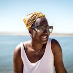 a black person smiling and looking into the distance. The sea is behind them. They have big glasses and wear a white tank top. 