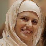 an olive-skinned woman smiles at the camera. She wears a loose cream headscarf. 