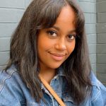 A close-up image of a black woman who smiles at the camera. She has medium length dark brown hair. She wears a blue denim jacket and a light brown shoulder bag. Behind Sheila is a grey tiled wall. 