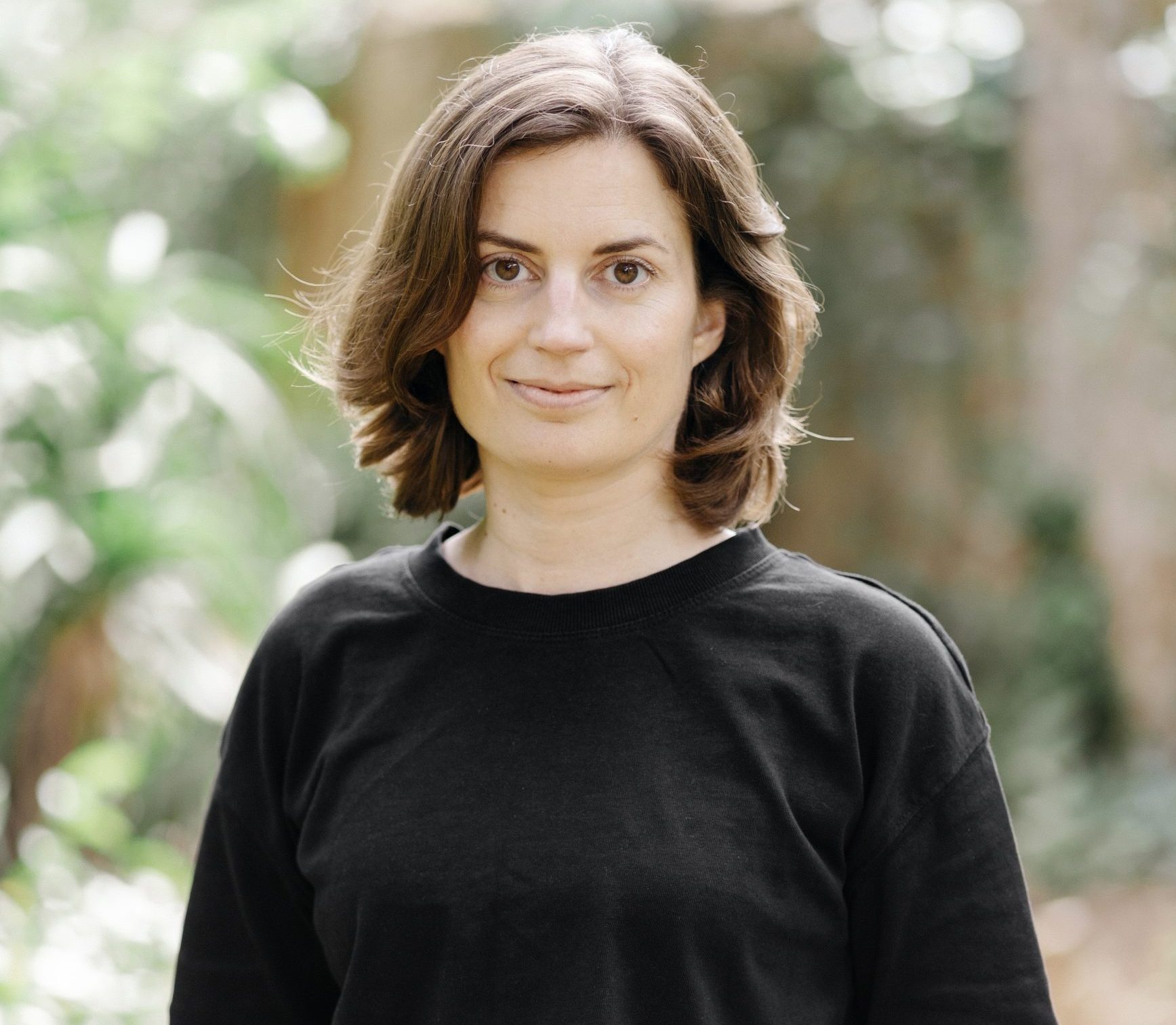 An image of Ana Fletcher, stood outside in a garden. She smiles, with one hand in her pocket. She is a white woman with short, shoulder-length hair brown hair. She wears a black t-shirt and black trousers.