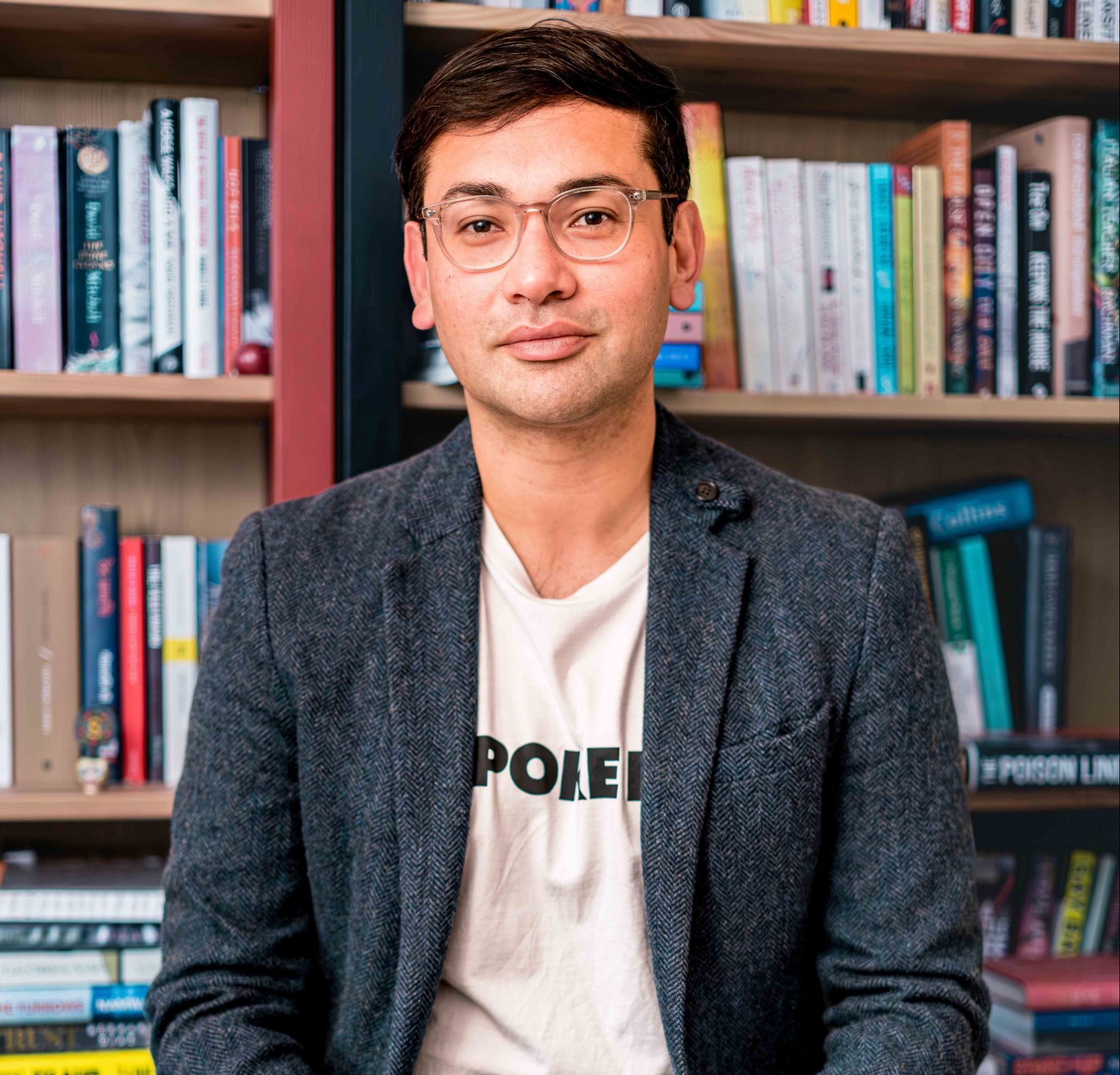 An image of Santanu, sitting down and facing the camera. Santanu is a light-skinned brown man with short dark hair, who wears glasses. He has a dark grey blazer and a white t-shirt beneath, and wears blue jeans. Behind him are bookshelves.