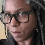 A close-up photo of Pam Williams, a black woman with long black and grey dread locks. She looks at the camera. She wears big black glasses and a black collared shirt.