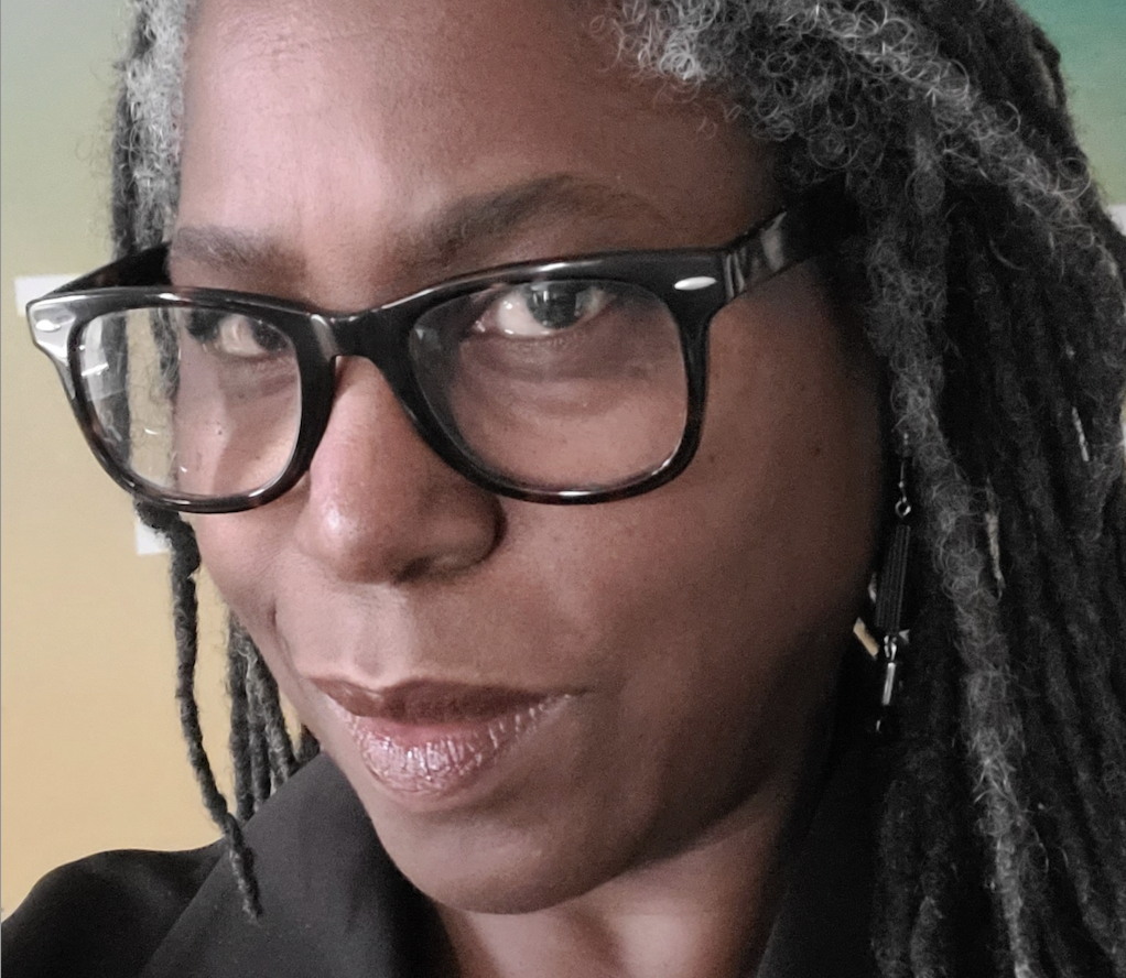 A close-up photo of Pam Williams, a black woman with long black and grey dread locks. She looks at the camera. She wears big black glasses and a black collared shirt.