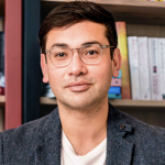 An image of Santanu, sitting down and facing the camera. Santanu is a light-skinned brown man with short dark hair, who wears glasses. He has a dark grey blazer and a white t-shirt beneath, and wears blue jeans. Behind him are bookshelves.
