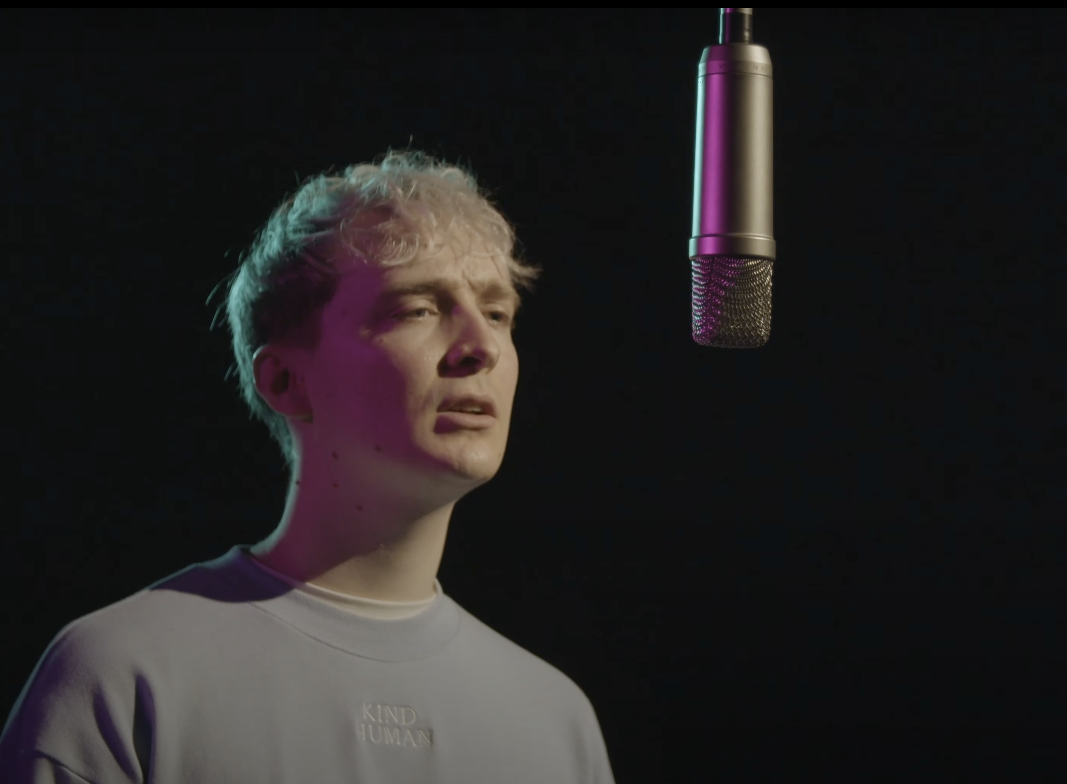 A light-skinned white person with blonde hair speaking into a microphone during a performance. The background is black, and they are wearing a light-blue jumper that reads 'Kind Human' in its centre. They are looking into the distance.