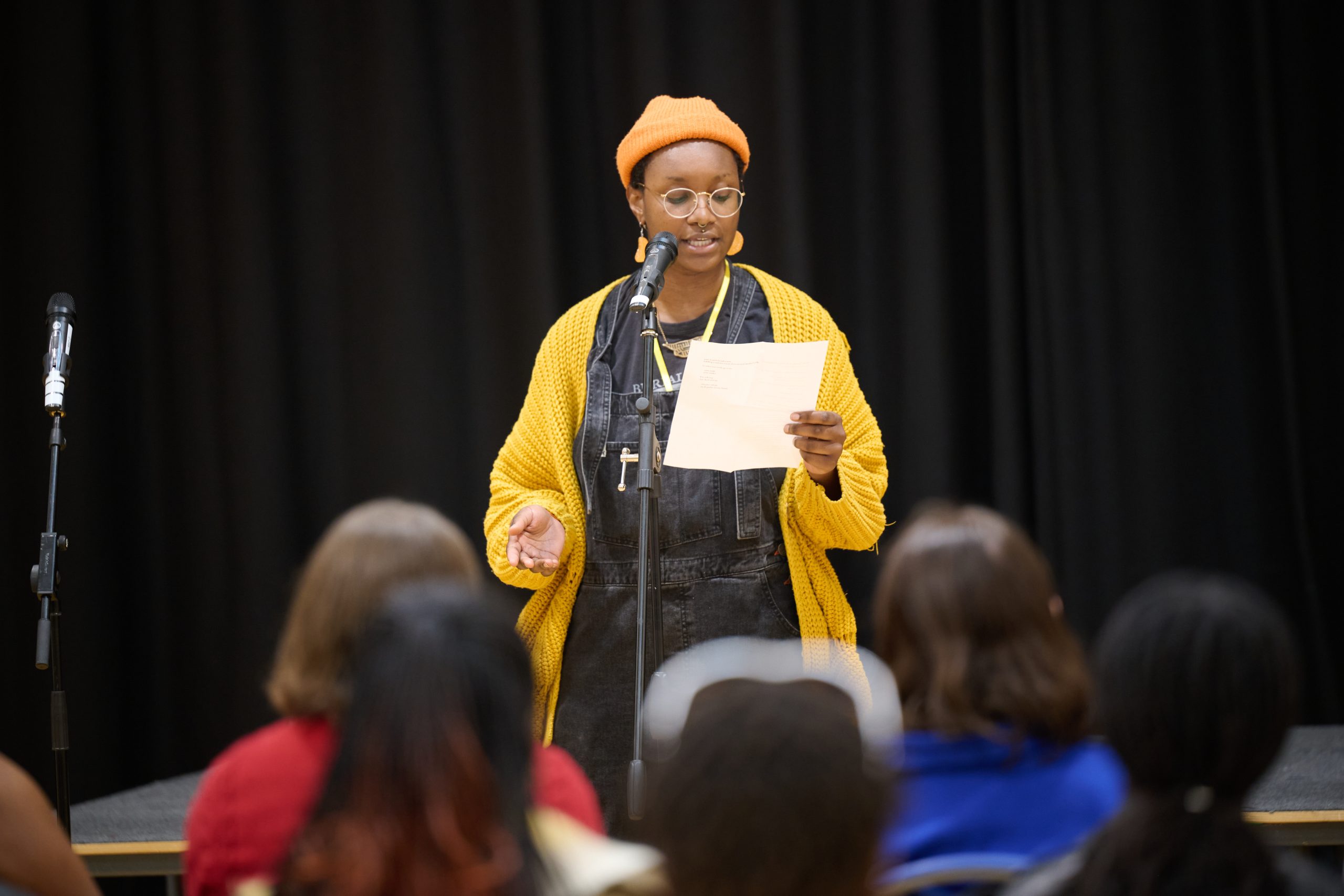 A young woman stands in front of an audience and a microphone, reading from a piece of paper she holds in one hand.