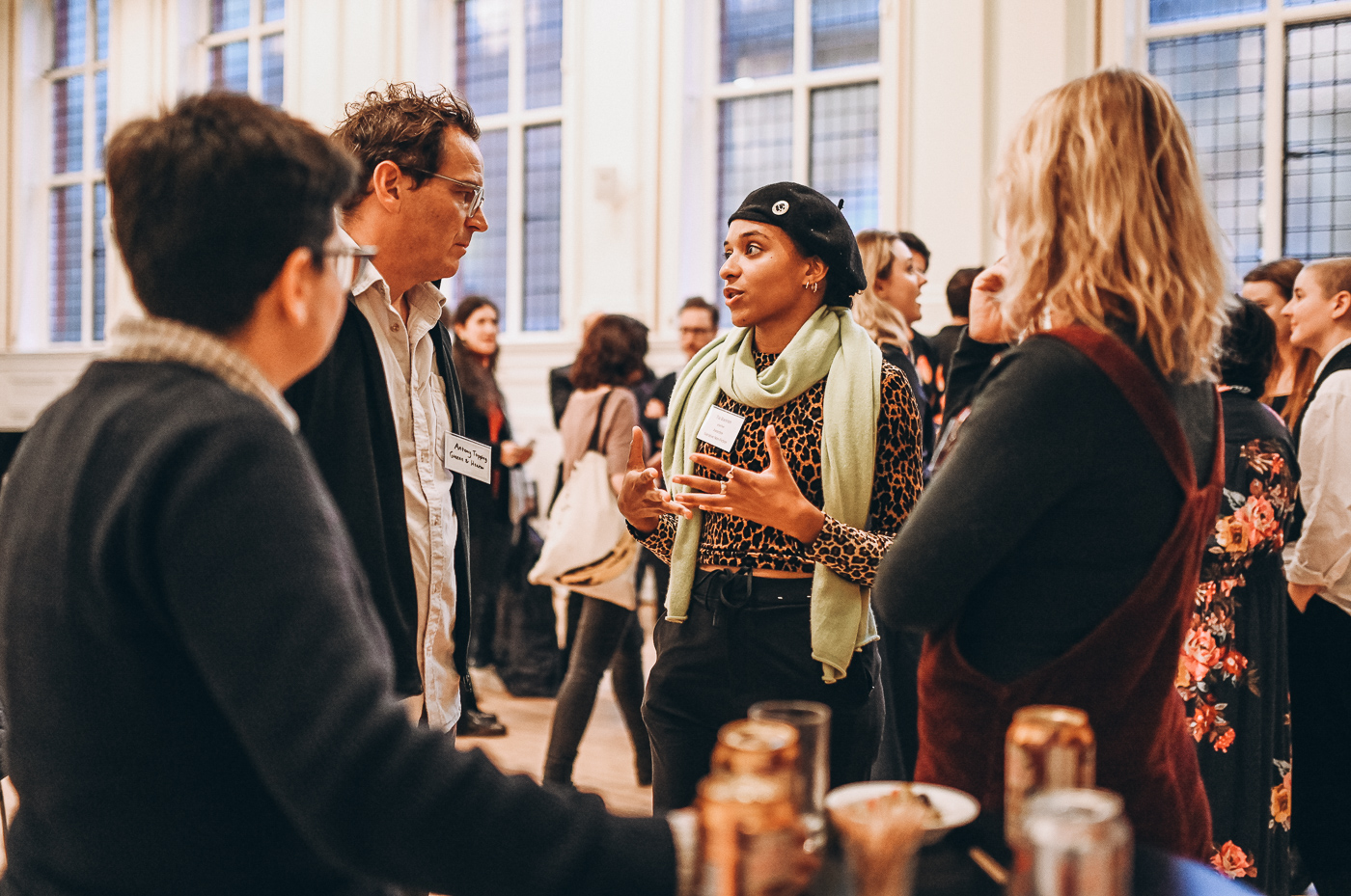 A group of people engage in a lively discussion at an event, with a focus on a woman wearing a green scarf and black beret as she speaks animatedly. The setting features large windows and a casual networking atmosphere.