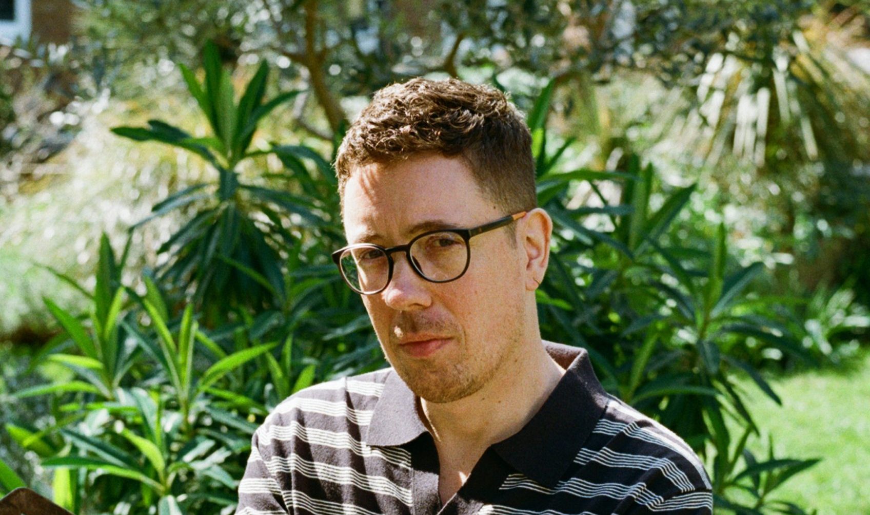 A light-skinned male with dark hair, arm tattoos and spectacles sits on a bench with his legs crossed. He is wearing dark trousers and a striped polo shirt. In the background are trees and plants.