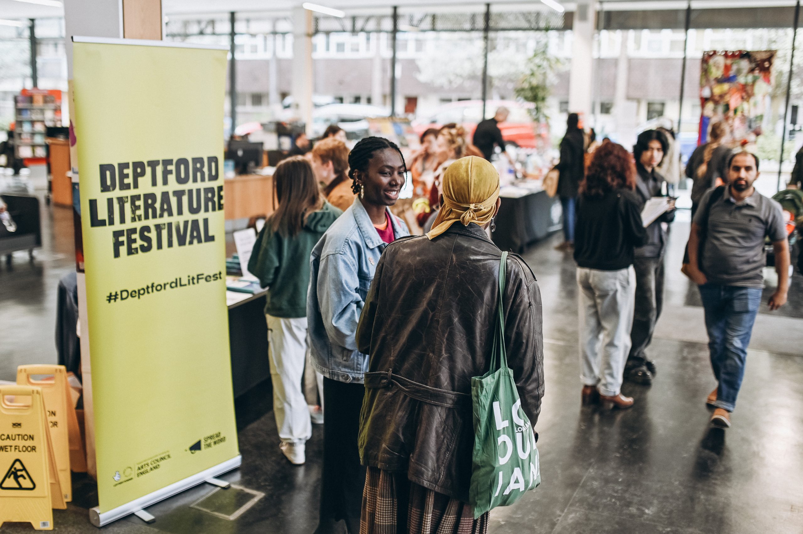 People interact and browse books at an indoor event for the Deptford Literature Festival, indicated by a yellow banner and visible book displays.