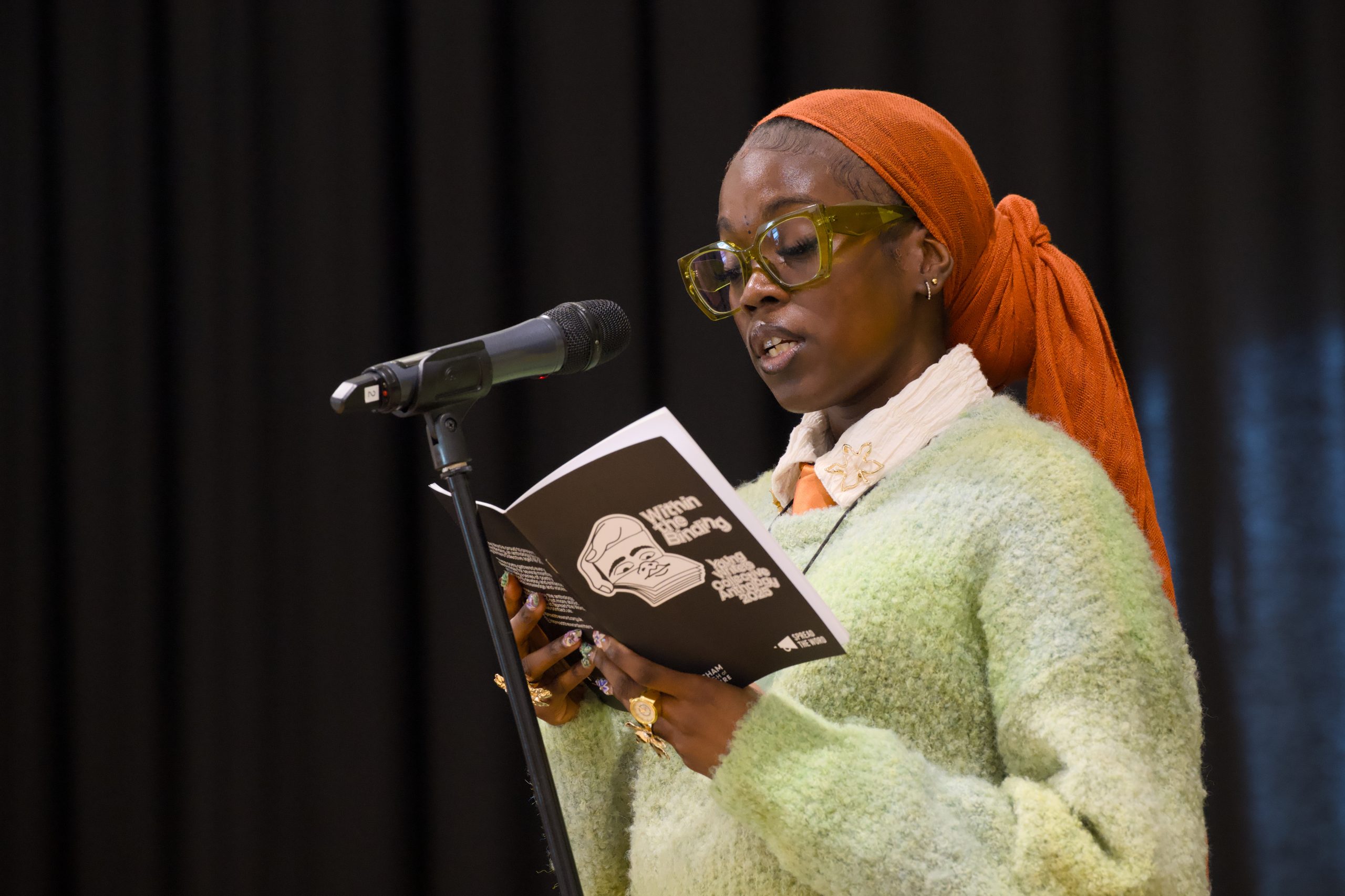 A person in glasses and an orange headscarf reads from a book at a microphone. They wear a green sweater and exude a focused, thoughtful expression.
