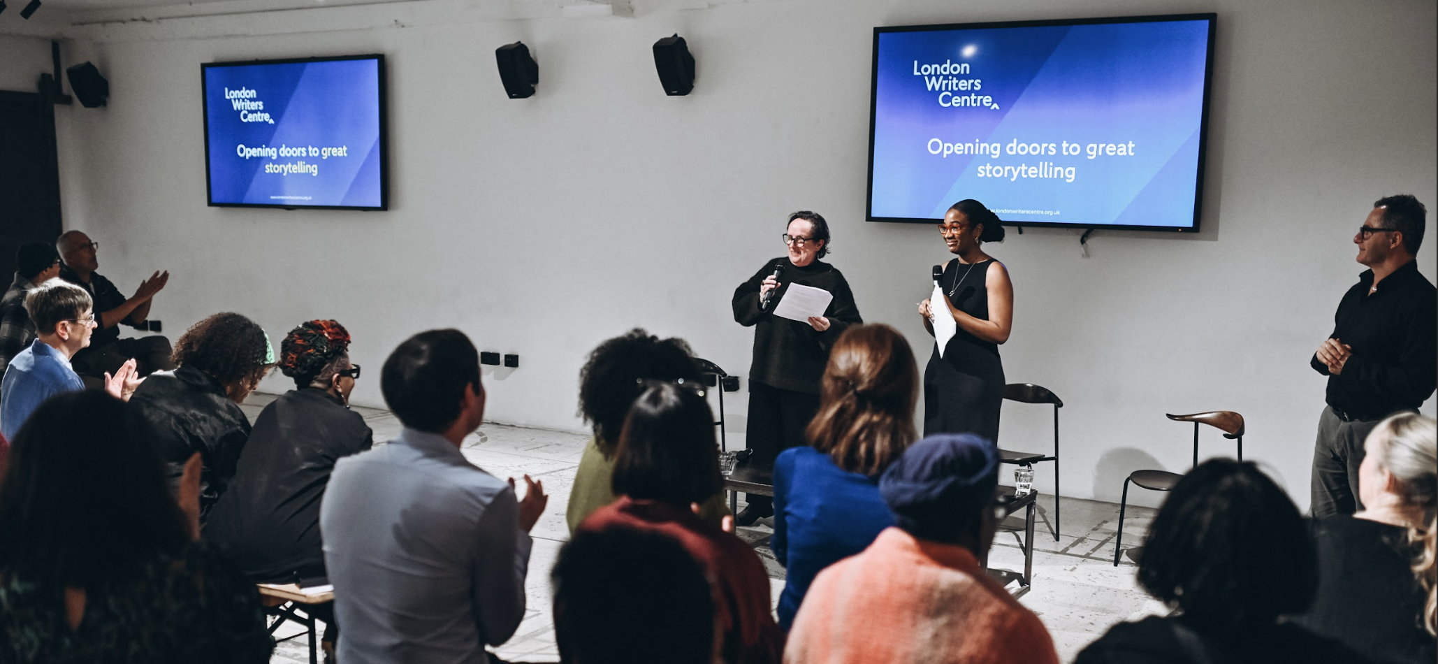 A warm, welcoming scene of a small audience applauding as two presenters stand at the front of a bright room, smiling beside a large screen displaying event graphics.