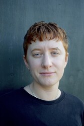 A person with short reddish hair and a slight smile looks directly at the camera against a plain, dark background, conveying a calm and approachable demeanor.